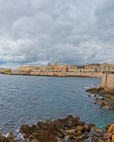 A coastal view of a historic walled city with ochre and yellow buildings lining the seafront under a cloudy sky, featuring a stone breakwater and rocky shoreline in the foreground.
