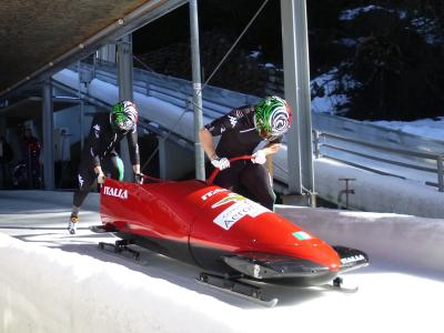 Two athletes in black racing suits and green-and-white helmets are pushing a red two-man bobsled labeled "ITALIA" and "Centro Sportivo Aeronautica Militare" on an outdoor ice track with snow banks and metal support structures.