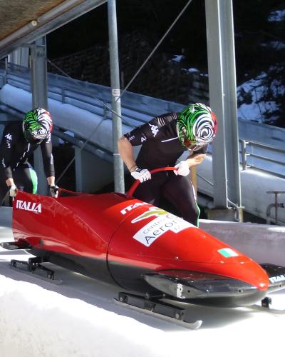Two athletes in black racing suits and green-and-white helmets are pushing a red two-man bobsled labeled "ITALIA" and "Centro Sportivo Aeronautica Militare" on an outdoor ice track with snow banks and metal support structures.