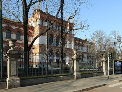 The image shows a large, ornate red-brick and white-stone building with Gothic Revival architectural features, including pointed arches and decorative pinnacles, viewed from behind a black metal fence and stone pillars on a city street.