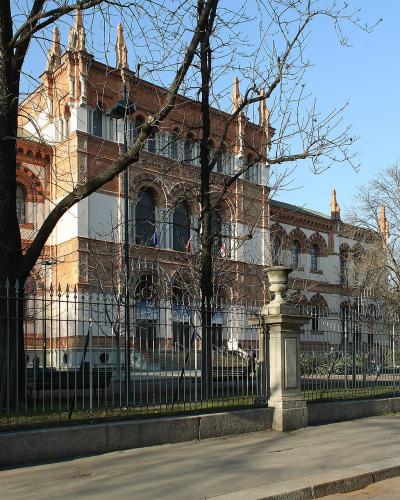 The image shows a large, ornate red-brick and white-stone building with Gothic Revival architectural features, including pointed arches and decorative pinnacles, viewed from behind a black metal fence and stone pillars on a city street.