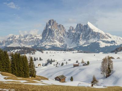 A panoramic winter landscape showing a snow-covered alpine meadow with scattered wooden huts, coniferous trees, and a prominent jagged mountain peak in the background under a partly cloudy sky.