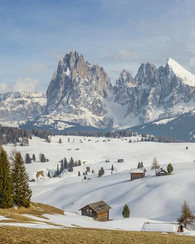 A panoramic winter landscape showing a snow-covered alpine meadow with scattered wooden huts, coniferous trees, and a prominent jagged mountain peak in the background under a partly cloudy sky.