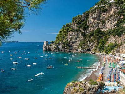 A scenic coastal view of a pebble beach lined with colorful umbrellas and sun loungers, backed by steep cliffs with buildings and a prominent stone tower (Castello di Fornillo) at the head of the bay, under a clear blue sky with numerous boats anc