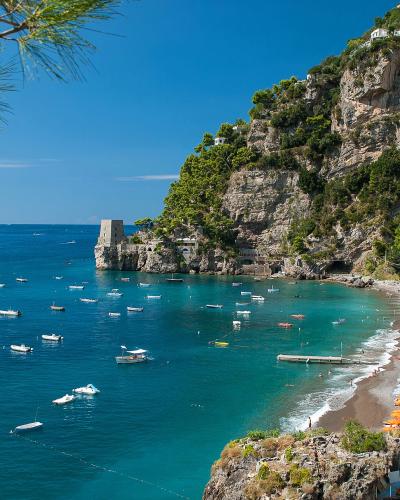 A scenic coastal view of a pebble beach lined with colorful umbrellas and sun loungers, backed by steep cliffs with buildings and a prominent stone tower (Castello di Fornillo) at the head of the bay, under a clear blue sky with numerous boats anc