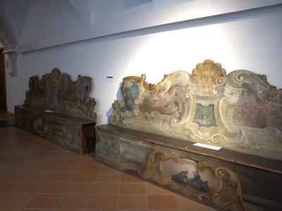 The image shows two large, ornate, painted wooden benches or stalls with elaborate baroque-style carvings and frescoes, displayed indoors on a terracotta tile floor under artificial lighting.