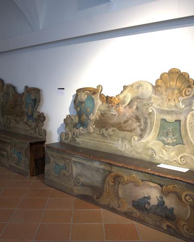 The image shows two large, ornate, painted wooden benches or stalls with elaborate baroque-style carvings and frescoes, displayed indoors on a terracotta tile floor under artificial lighting.