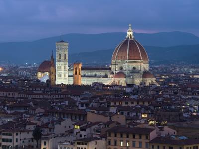 A twilight view of the Florence Cathedral complex, featuring Brunelleschi's dome, Giotto's Campanile, and the Baptistery, illuminated against a backdrop of hills and the city's rooftops.