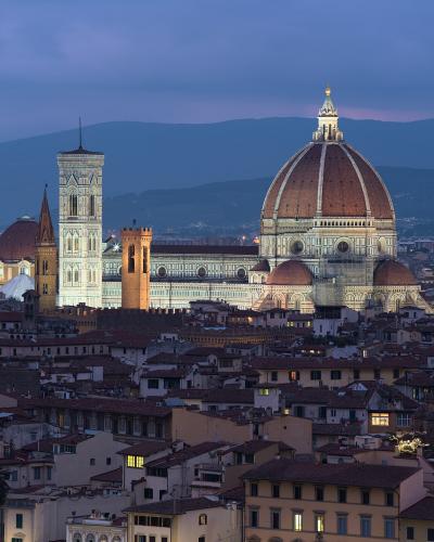 A twilight view of the Florence Cathedral complex, featuring Brunelleschi's dome, Giotto's Campanile, and the Baptistery, illuminated against a backdrop of hills and the city's rooftops.