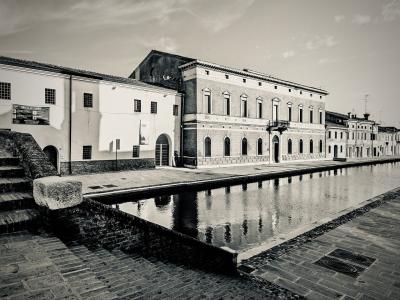 A black and white photograph showing a large, multi-story brick building with arched windows on the ground floor and rectangular windows above, situated next to a canal or waterway in an Italian town setting.