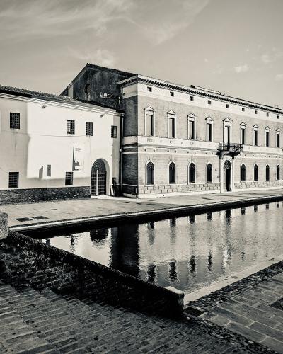 A black and white photograph showing a large, multi-story brick building with arched windows on the ground floor and rectangular windows above, situated next to a canal or waterway in an Italian town setting.