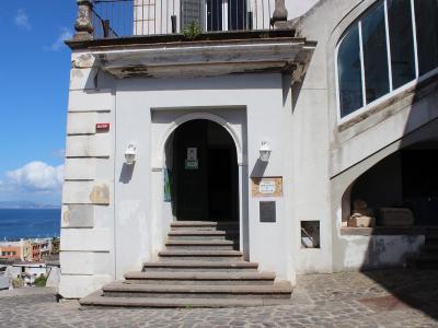 The image shows the entrance to a white building with a stone staircase leading up to an arched doorway; a sign on the wall to the right of the door reads "MUSEO ARCHEOLOGICO" and "ISCHIA", and the sea is visible in the background.