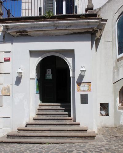 The image shows the entrance to a white building with a stone staircase leading up to an arched doorway; a sign on the wall to the right of the door reads "MUSEO ARCHEOLOGICO" and "ISCHIA", and the sea is visible in the background.