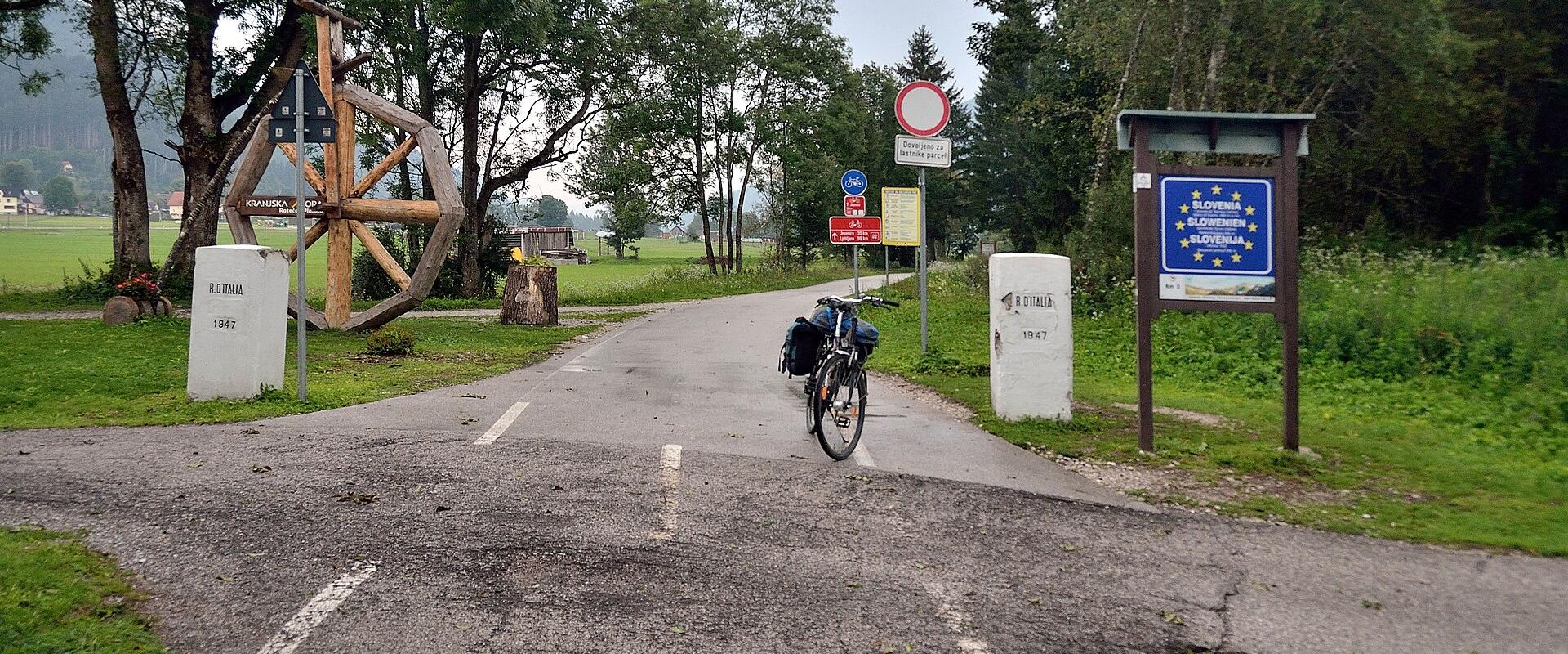 A paved road at the Italy-Slovenia border crossing near Rateče, marked by white stone pillars inscribed "ITALIA 1947" and "SLOVENIJA 1947", a cyclist with touring gear, and a blue sign displaying the Slovenian flag and EU stars.