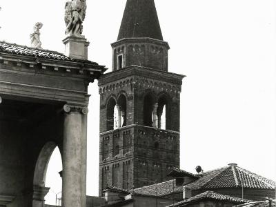 A black-and-white photograph showing the brick bell tower (campanile) of a church with a pointed spire, viewed from behind a classical-style loggia featuring columns and statues on its roofline.