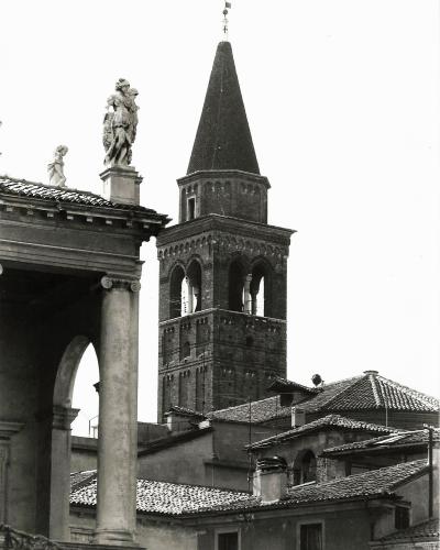 A black-and-white photograph showing the brick bell tower (campanile) of a church with a pointed spire, viewed from behind a classical-style loggia featuring columns and statues on its roofline.