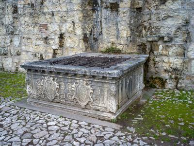 A stone cistern or water basin with ornate carvings and a metal grate, set against a rough stone wall on a cobblestone path.