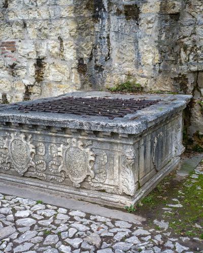 A stone cistern or water basin with ornate carvings and a metal grate, set against a rough stone wall on a cobblestone path.