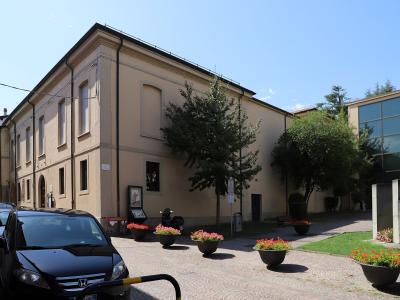 A beige, two-story building with arched windows and a flat roofline, situated on a paved plaza with flower planters, trees, parked cars, and a modern glass building to the right; signage includes an information kiosk and a small sign near the entr