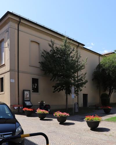 A beige, two-story building with arched windows and a flat roofline, situated on a paved plaza with flower planters, trees, parked cars, and a modern glass building to the right; signage includes an information kiosk and a small sign near the entr