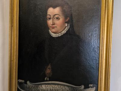 A framed oil painting of a woman in period attire, identified by an inscription at the bottom as "LIONORA GONZAGA VXOR FRANCISCI MARIAE I. V.D.", hanging on a white wall inside a building.