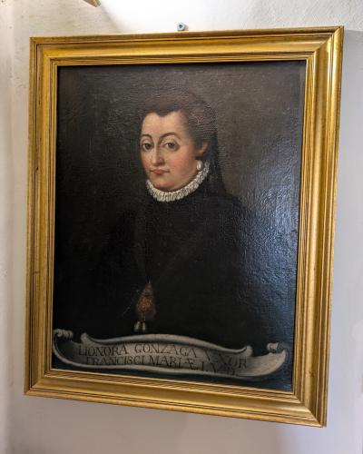 A framed oil painting of a woman in period attire, identified by an inscription at the bottom as "LIONORA GONZAGA VXOR FRANCISCI MARIAE I. V.D.", hanging on a white wall inside a building.