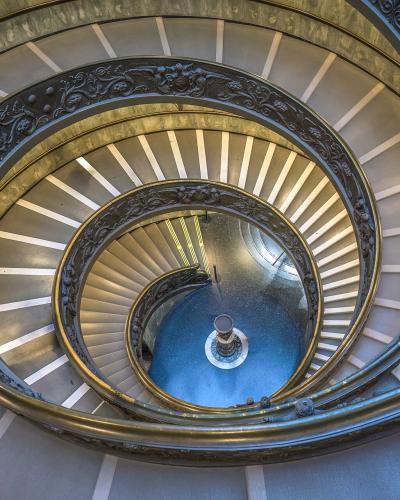 A top-down view of a double-helix spiral staircase with ornate dark metal railings and light-colored steps, located indoors.
