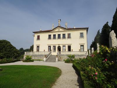 A cream-colored two-story villa with a central pediment topped by statues, a balustraded terrace with stairs, gravel paths, manicured lawns, and rose bushes in the foreground under a blue sky.