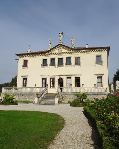 A cream-colored two-story villa with a central pediment topped by statues, a balustraded terrace with stairs, gravel paths, manicured lawns, and rose bushes in the foreground under a blue sky.