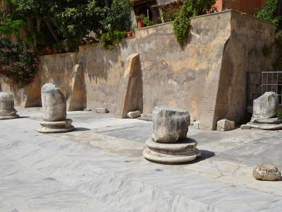 The image shows a paved courtyard or plaza with several ancient stone column bases and fragments, set against a weathered retaining wall with vegetation and potted plants above.