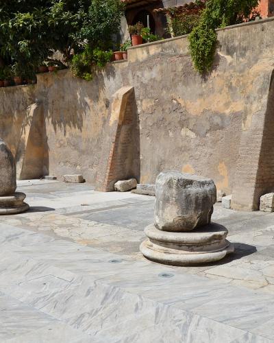 The image shows a paved courtyard or plaza with several ancient stone column bases and fragments, set against a weathered retaining wall with vegetation and potted plants above.