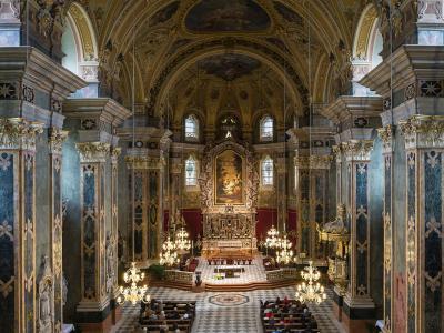The image shows the ornate Baroque interior of a cathedral, featuring a high vaulted ceiling with frescoes, large marble-clad columns with gold capitals, a gilded high altar with a central painting, and rows of pews with congregants.