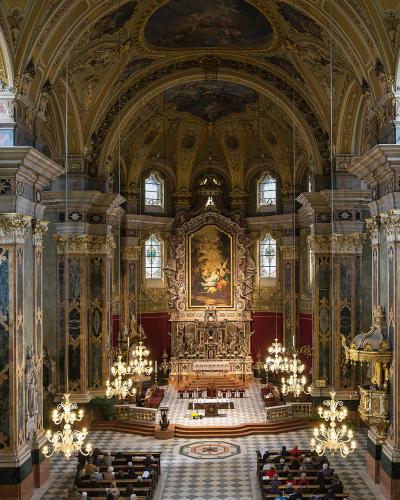 The image shows the ornate Baroque interior of a cathedral, featuring a high vaulted ceiling with frescoes, large marble-clad columns with gold capitals, a gilded high altar with a central painting, and rows of pews with congregants.