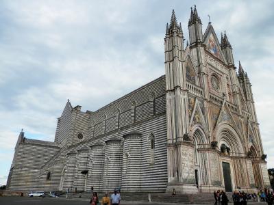 A low-angle photograph of a large Gothic cathedral with distinctive black-and-white horizontal striped masonry on its side walls and an ornate, multi-tiered facade featuring mosaics, statues, and pointed spires, with tourists visible at its base.