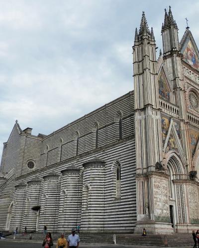 A low-angle photograph of a large Gothic cathedral with distinctive black-and-white horizontal striped masonry on its side walls and an ornate, multi-tiered facade featuring mosaics, statues, and pointed spires, with tourists visible at its base.