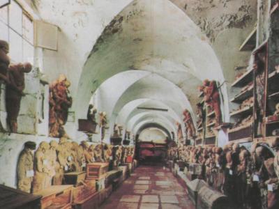 A long, arched underground corridor lined with rows of mummified human figures and skeletal remains arranged on shelves and platforms along both walls.