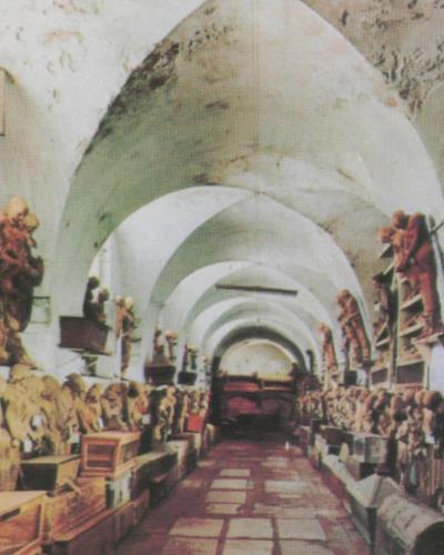 A long, arched underground corridor lined with rows of mummified human figures and skeletal remains arranged on shelves and platforms along both walls.