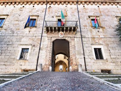 A low-angle photograph of a large, historic stone building facade featuring a central arched gateway, a small balcony with an Italian flag, and a cobblestone ramp leading up to it.