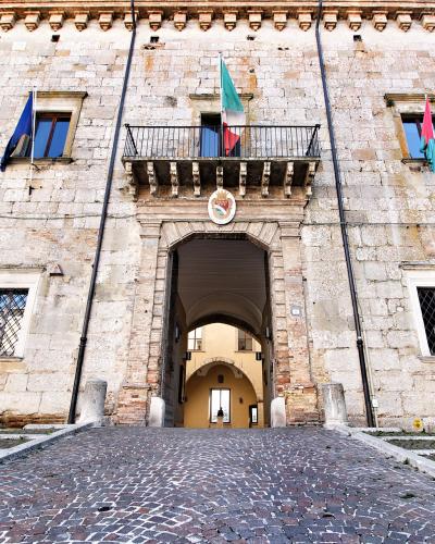 A low-angle photograph of a large, historic stone building facade featuring a central arched gateway, a small balcony with an Italian flag, and a cobblestone ramp leading up to it.