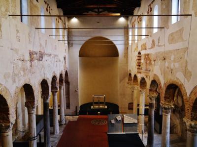 The image shows the interior nave of a historic church with exposed brick arches, ancient columns, peeling plaster walls revealing frescoes, and a red carpet leading to an apse; modern display cases and lighting fixtures are present, indicating it