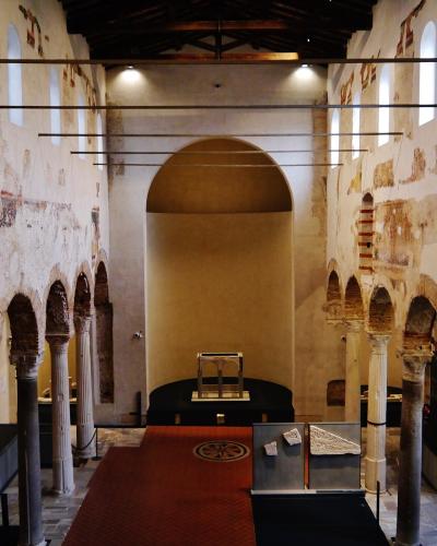 The image shows the interior nave of a historic church with exposed brick arches, ancient columns, peeling plaster walls revealing frescoes, and a red carpet leading to an apse; modern display cases and lighting fixtures are present, indicating it