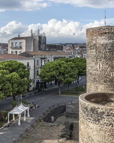 A high-angle view from atop a stone fortress wall showing a cylindrical tower, a brick parapet, and a street lined with green trees leading toward city buildings under a cloudy sky.