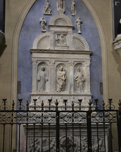A detailed white marble funerary monument or tomb effigy set within a blue-painted arched niche, featuring multiple sculpted figures and enclosed by a black wrought-iron fence.