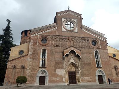 A low-angle photograph of a large, historic brick cathedral facade featuring a central rose window, Gothic-style pointed arch doorways, and a prominent stepped gable under an overcast sky.