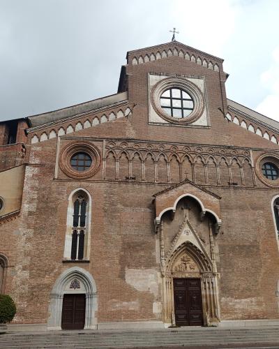 A low-angle photograph of a large, historic brick cathedral facade featuring a central rose window, Gothic-style pointed arch doorways, and a prominent stepped gable under an overcast sky.