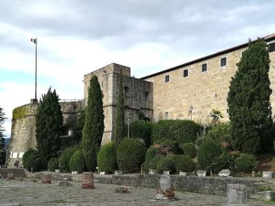 A stone fortress with a tall square tower, a long rectangular wing with small barred windows, a round bastion flying an Italian flag, and a foreground courtyard with scattered ancient stone fragments and cypress trees.