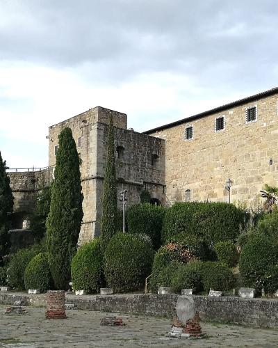 A stone fortress with a tall square tower, a long rectangular wing with small barred windows, a round bastion flying an Italian flag, and a foreground courtyard with scattered ancient stone fragments and cypress trees.