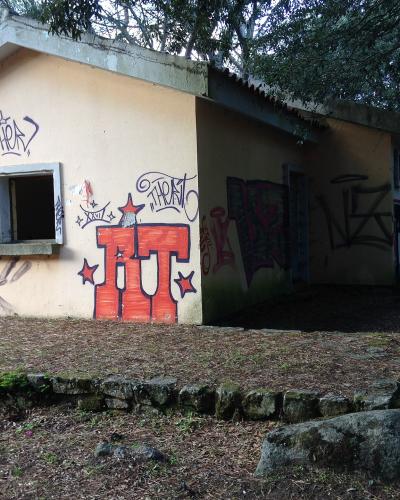 A small, single-story building with a flat roof and beige walls, heavily covered in graffiti (including large red letters "IT" and tags like "THOR"), situated on a leafy, wooded slope with stone retaining walls.