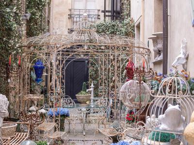 The image shows an outdoor display of ornate, antique-style metalwork (including large birdcages, furniture, and statues) and potted plants, likely the storefront of an antique or garden decor shop.