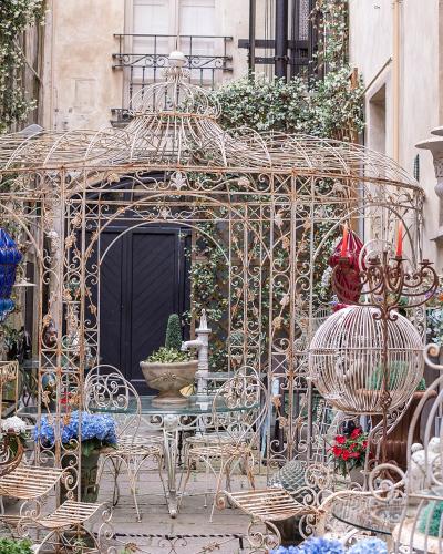 The image shows an outdoor display of ornate, antique-style metalwork (including large birdcages, furniture, and statues) and potted plants, likely the storefront of an antique or garden decor shop.